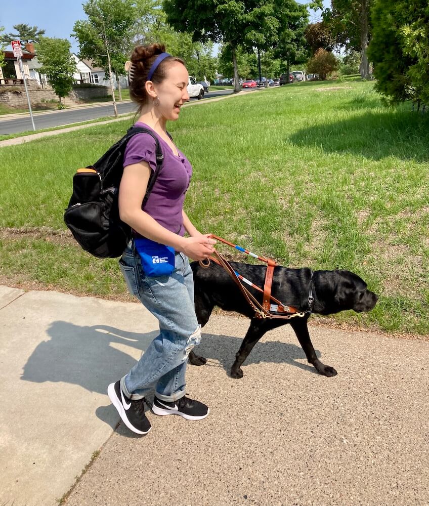 Rachel happily walks in the sun down a sidewalk with guide dog Chris