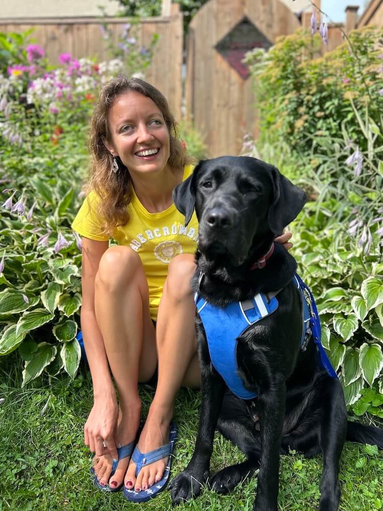 Weronika happily sits with black lab guide dog Reggae in at edge of a garden