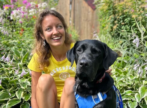 Weronika happily sits with black lab guide dog Reggae in at edge of a garden