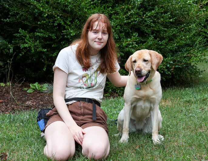 Raiser Aileen kneels in grass with hand of the back of released yellow Lab Paisley