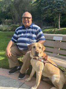 Latif sits with yellow Lab Guide dog Sugar on an outdoor bench in their team portrait