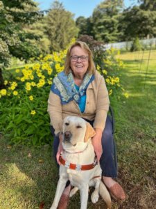 Angela sits in front of yellow flowers in a yard with yellow Lab guide dog Alyssa