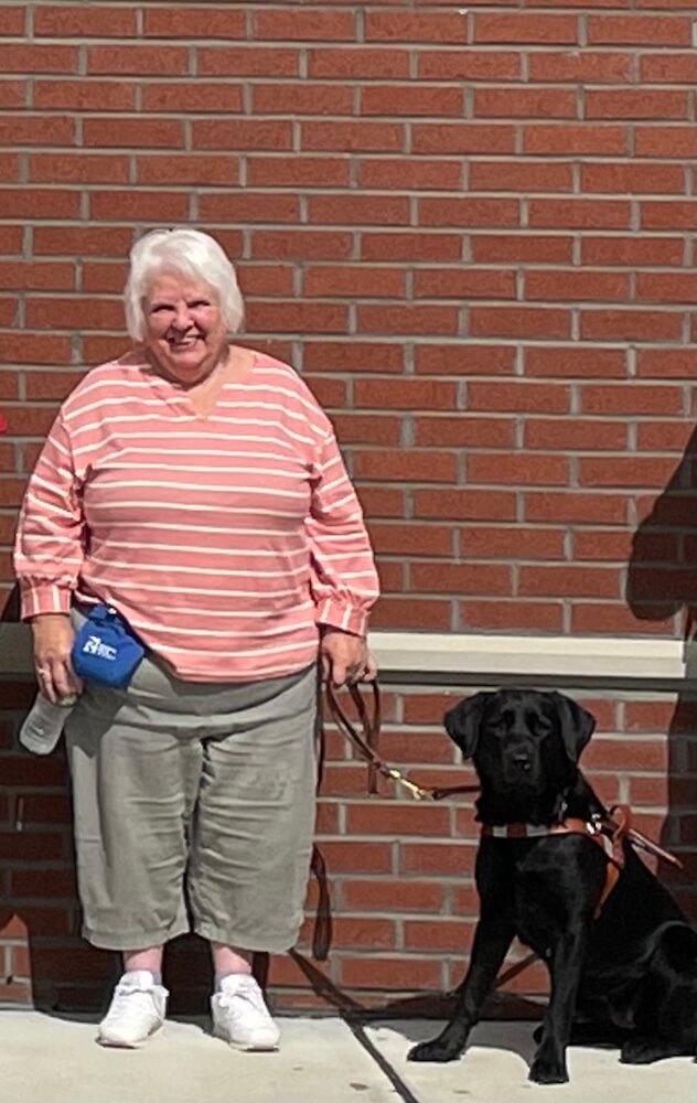 Cathy stands in front of brick wall smiling while holding Meg's leash
