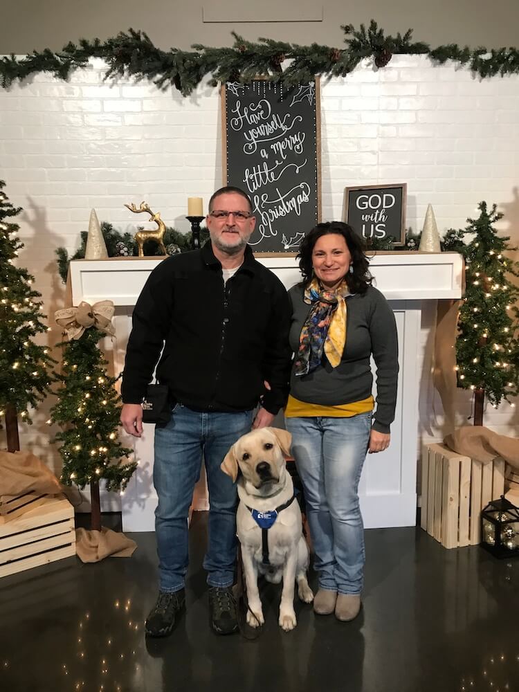 Pup on program Francisco poses with raisers in front of holiday decorated fireplace