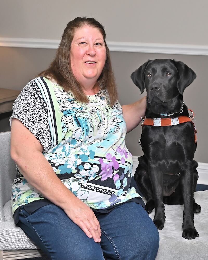 Gail sits next to black Lab guide dog Danielle for team portrait