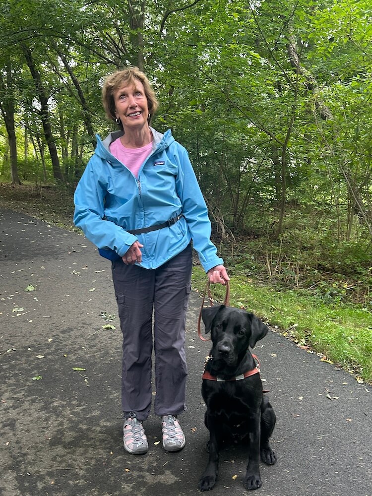 Joyce and black Lab guide Harry stand in middle of a wooded path