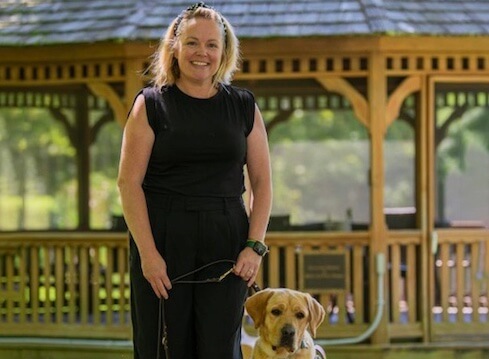 Kathleen O'Reilly, Planned Giving, with yellow Lab guide dog in harness stands near nature path gazebo smiling in long black dress