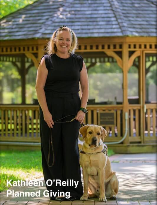 Kathleen O'Reilly, Planned Giving, with yellow Lab guide dog in harness stands near nature path gazebo smiling in long black dress