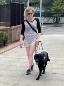Margot and black Lab guide dog Jennifer walking down a wide sidewalk