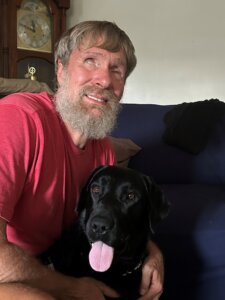 Teddy has arm around black Lab guide Dan while sitting on a couch for team portrait