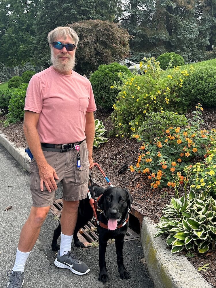 Teddy stands with guide Dan at a raised curb with colorful flowers and plantings