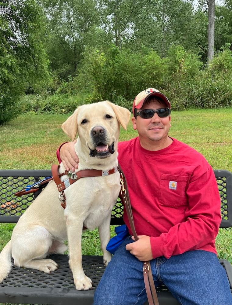 Travis sits happily on an outdoor bench with arm around yellow lab guide dog Francisco, also smiling