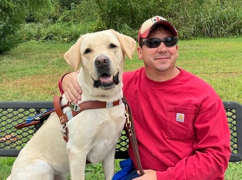 Travis sits happily on an outdoor bench with arm around yellow lab guide dog Francisco, also smiling