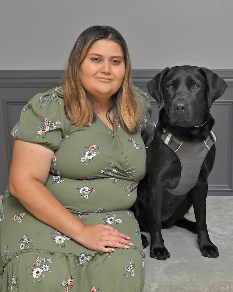 Aida sits on a gray bench with black Lab guide dog Bunnie next to her for team portrait