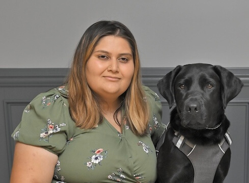 Aida sits on a gray bench with black Lab guide dog Bunnie next to her for team portrait
