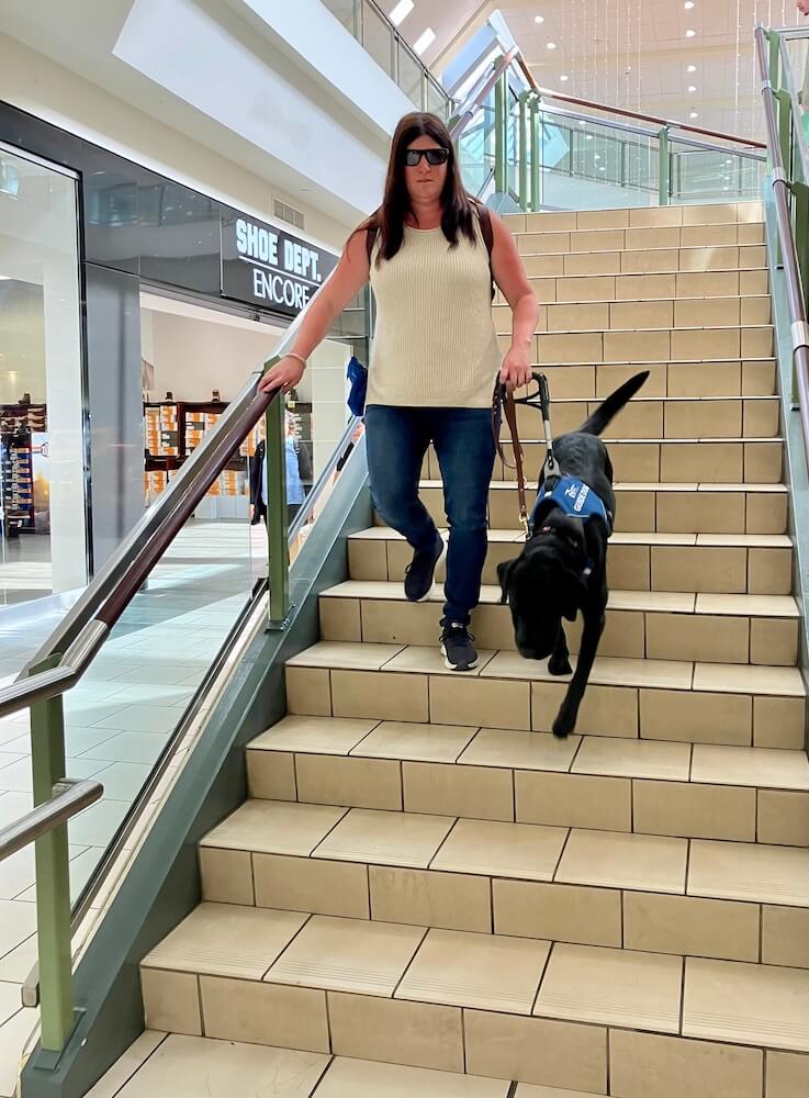 Kayla and black lab guide dog Cayenne walk safely down mall staircase