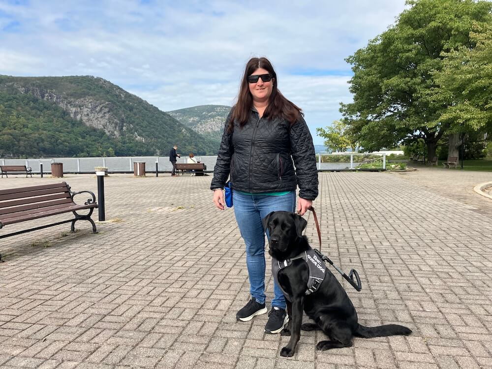 Kayla stands with guide dog Cayenne on brick pavement against backdrop of mountains