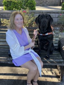 Linda sits on an outdoor bench with black Lab guide dog Edamame for team portrait