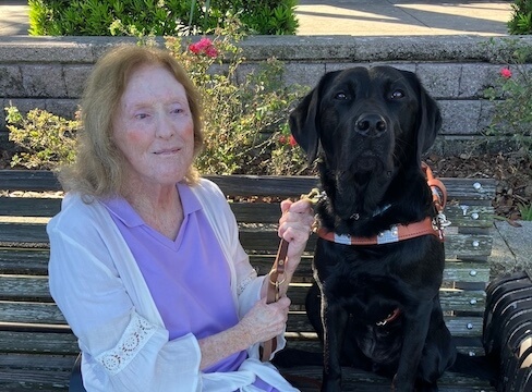 Linda sits on an outdoor bench with black Lab guide dog Edamame for team portrait