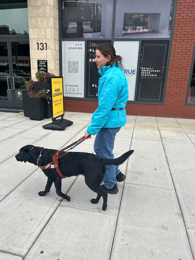 Candice and black Lab guide Charlotte walk down a wide sidewalk  in an urban area