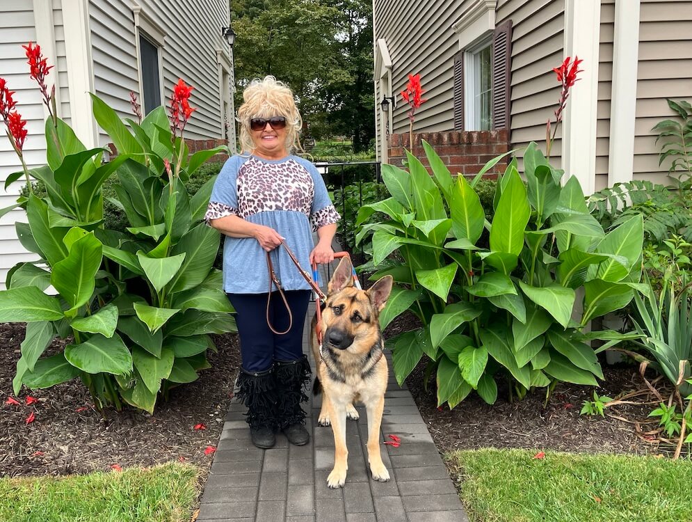 Deborrah and her German Shepherd guide Dixie stand happily between 2 clusters of foliage with tall red flowers