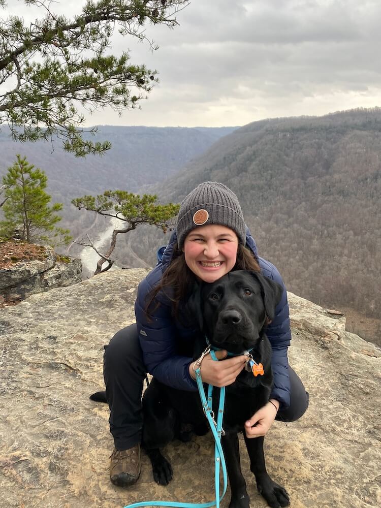 Black Lab Elliott on rocky ledge of a tall mountain overlooking a river valley with raiser Jenevieve
