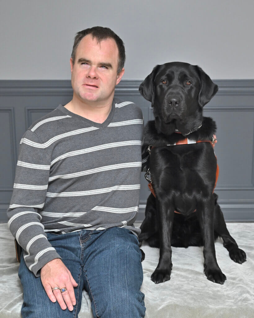 Michael and black Lab guide dog Emerson sit together for team portrait against gray background