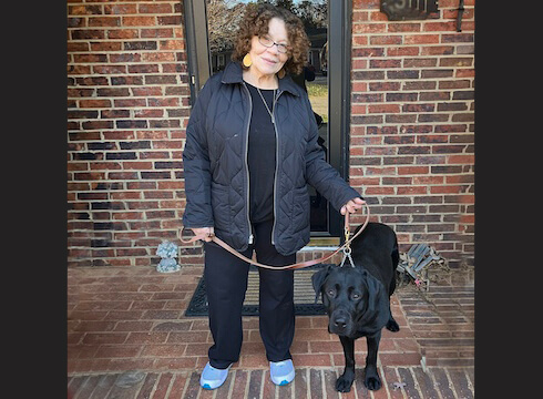Sandra and black Lab guide dog Elliott stand at entrance of a brick building looking at the camera