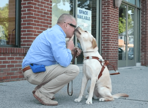 David squats and receives a kiss from yellow Lab guide dog Tessi with brick storefront behind them