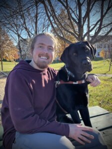 Joshua sits happily on an outdoor bench with black lab guide dog Quinn