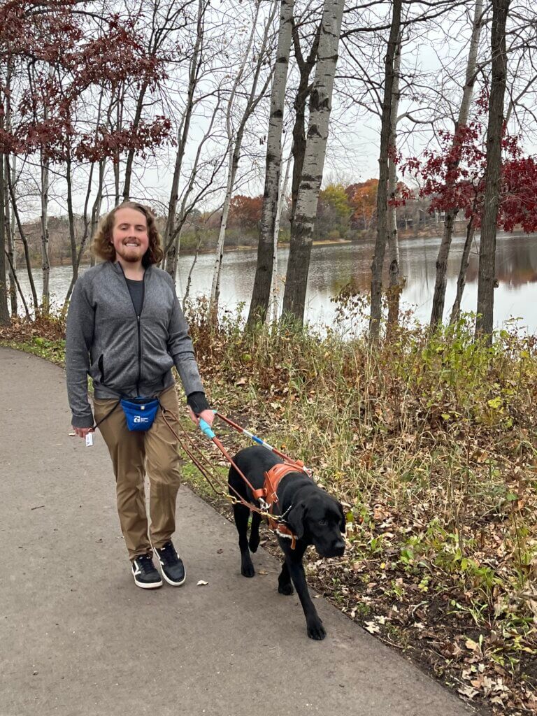 Josh walks safely down a paved path near a lake with guide dog Quinn