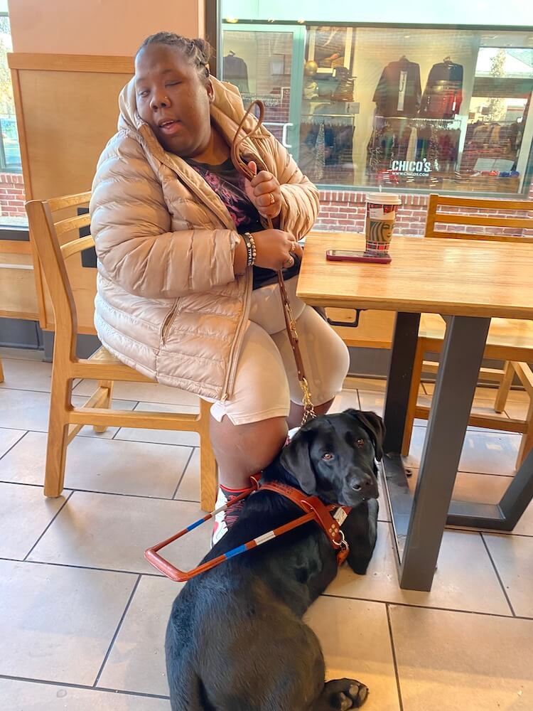 Dazia sits in a coffee shop and guide dog Casey lies quietly on the floor beneath the table