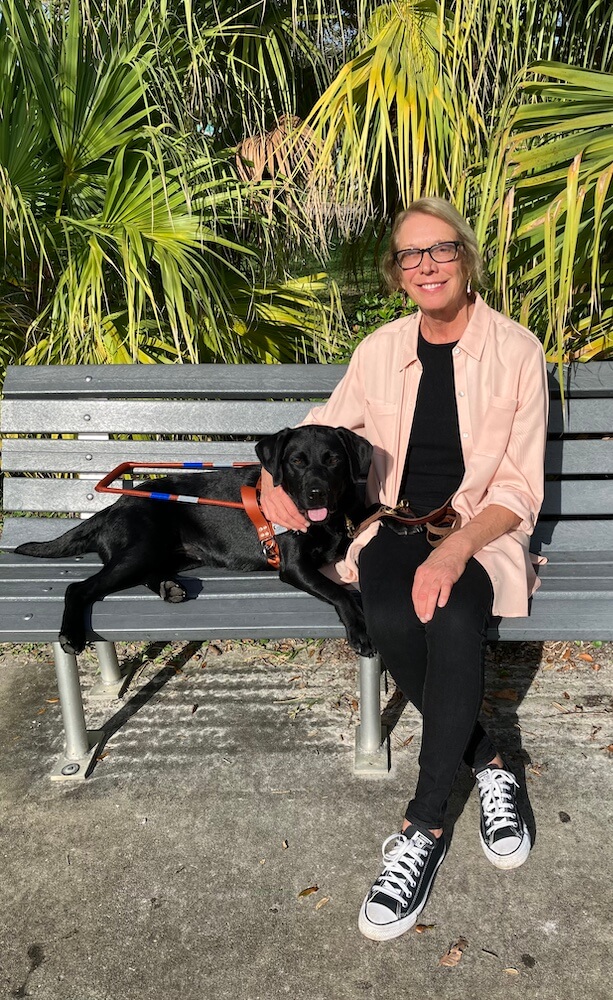 Black Lab guide dog Hildy reclines with her head next to Deni as they sit on an outdoor bench.
