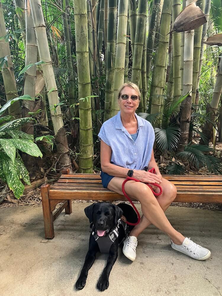 Deni sits on a bench in front of tropical trees and greenery with Hildy at her feet