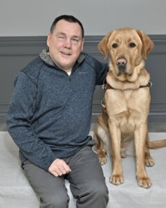 Andrew is next to yellow Lab guide dog  Zeb as they sit for their team portrait