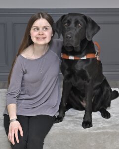 Caitlyn sits sits with black Lab guide dog Rory for their team portrait
