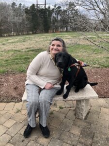 Deborah leans into black lab guide Scarlett while sitting on a stone outdoor bench