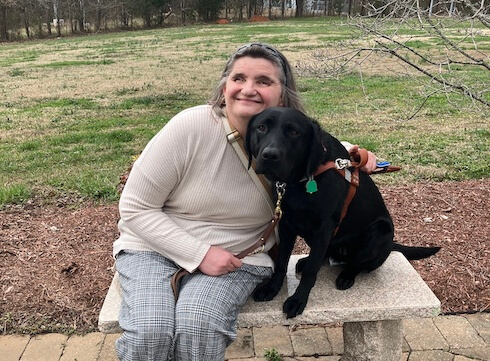 Deborah leans into black lab guide Scarlett while sitting on a stone outdoor bench