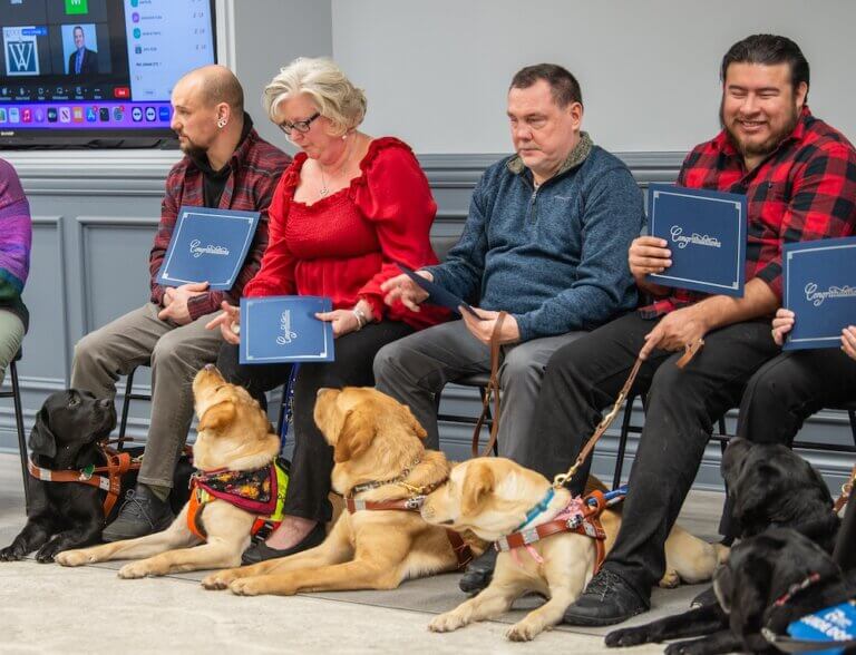 Four students sit at graduation holding their certificates while dogs lie at their feet