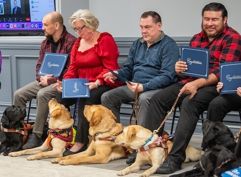 four graduates sit on folding chair holding their certificate with attentive guide dogs in a down looking up at them at graduation