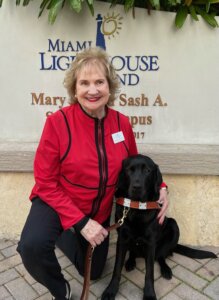 Virginia sits with black Lab guide Sama in front of a Miami Lighthouse sign