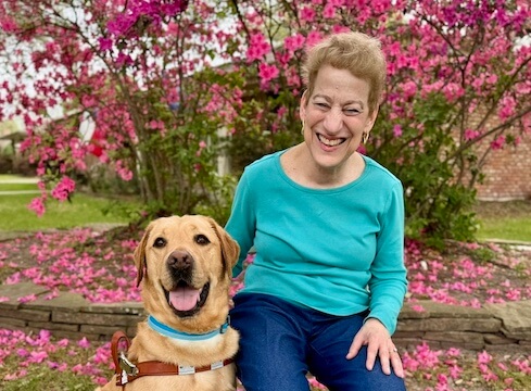 Anita sits in a chair in front of a bright pink azalea bush with yellow Lab Loki and both have very happy faces