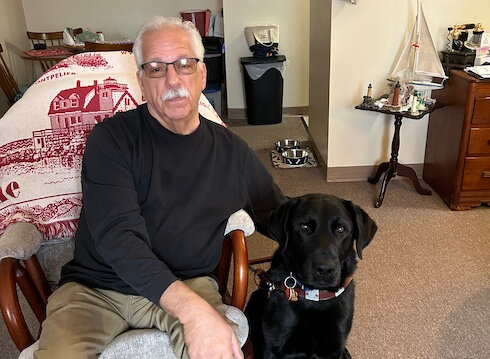 Craig sits in a chair in his home with guide Ephram sitting beside his legs.