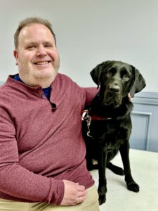 Gary happily sits beside black Lab guide dog Izzie for their graduate portrait
