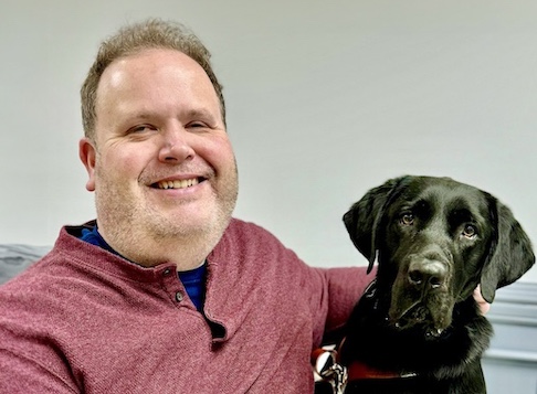 Gary happily sits beside black Lab guide dog Izzie for their graduate portrait