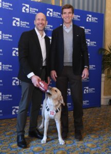 Eli Manning with Guiding Eyes CEO and President Thomas Panek with yellow guide dog, Ten, at The Penn Club January 2024. Ten who holds a football toy in his mouth.