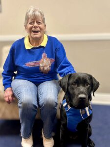 Karen happily sits in a chair with her black Lab guide dog Ohana sitting at her feet