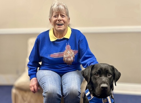 Karen happily sits in a chair with her black Lab guide dog Ohana sitting at her feet