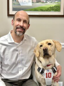 Thomas Panek sit with arm around guide dog Ten who wears a white harness with red 10 to mimic an NFL Giants jersey