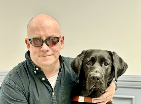 William has arm around neck of black Lab guide dog Quidditch as they sit for their team portrait